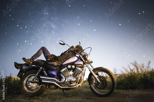 Young beautiful biker woman with motorcycle under stars of Miky Way galaxy . Female biker at night