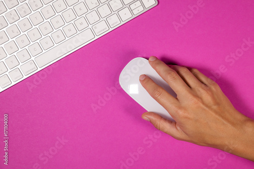 Computer keyboard and computer mouse on a pink background from above. Woman's hand touching the computer mouse