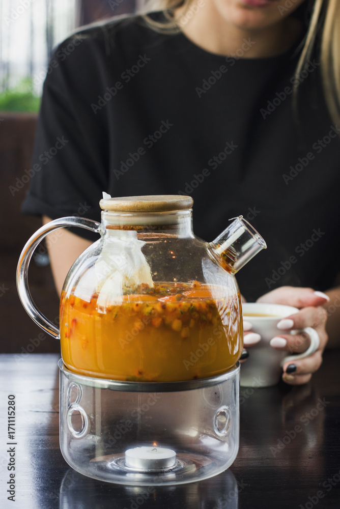 Sea buckthorn tea in the glass teapot.The young blonde girl drinking tea in the cafe on the background Sea buckthorn tea in the glass teapot.The young blonde girl drinking tea in the cafe on the background