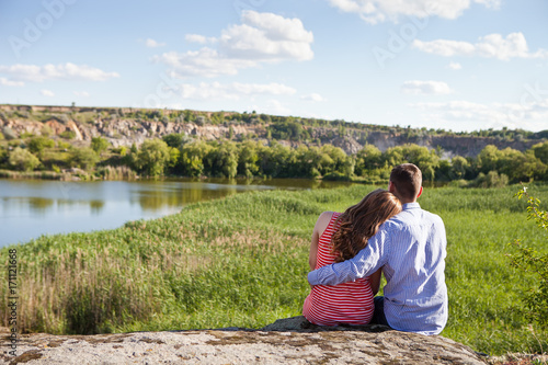 A young man and a girl are sitting with their backs to the camera on top of a hill in the background of a river landscape