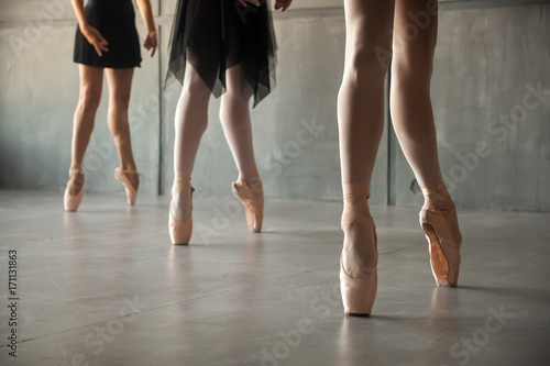 Canvas Print Close-up of the legs of three young  ballerinas in white pantyhose, black packs and pointes performing a dance in a dark studio