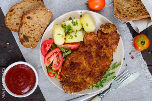 Golden schnitzel, boiled potatoes and salad with tomatoes on a plate on a dark wooden background.
