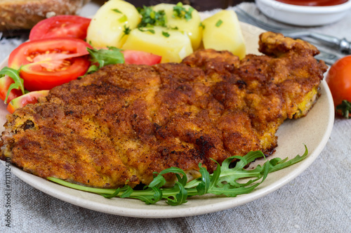 Golden schnitzel, boiled potatoes and salad with tomatoes on a plate on a dark wooden background.