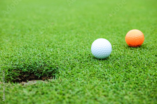 White and orange color golf ball on green near hole at the morning time