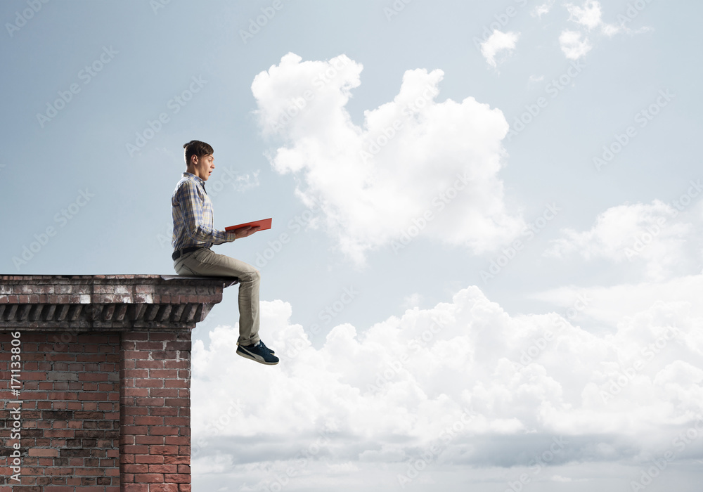 Man on roof edge reading book and cloudscape at background Man on roof edge reading book and cloudscape at background