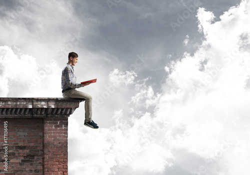 Man on roof edge reading book and cloudscape at background
