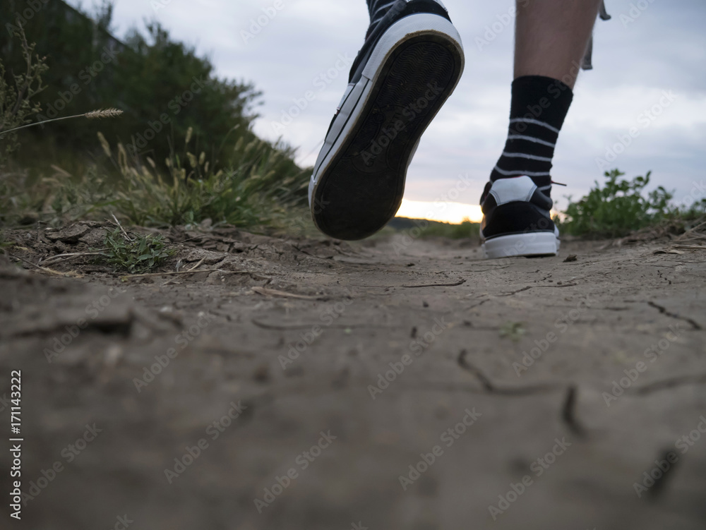 Man feet walking on dry dirt path in sunset and cloudy sky