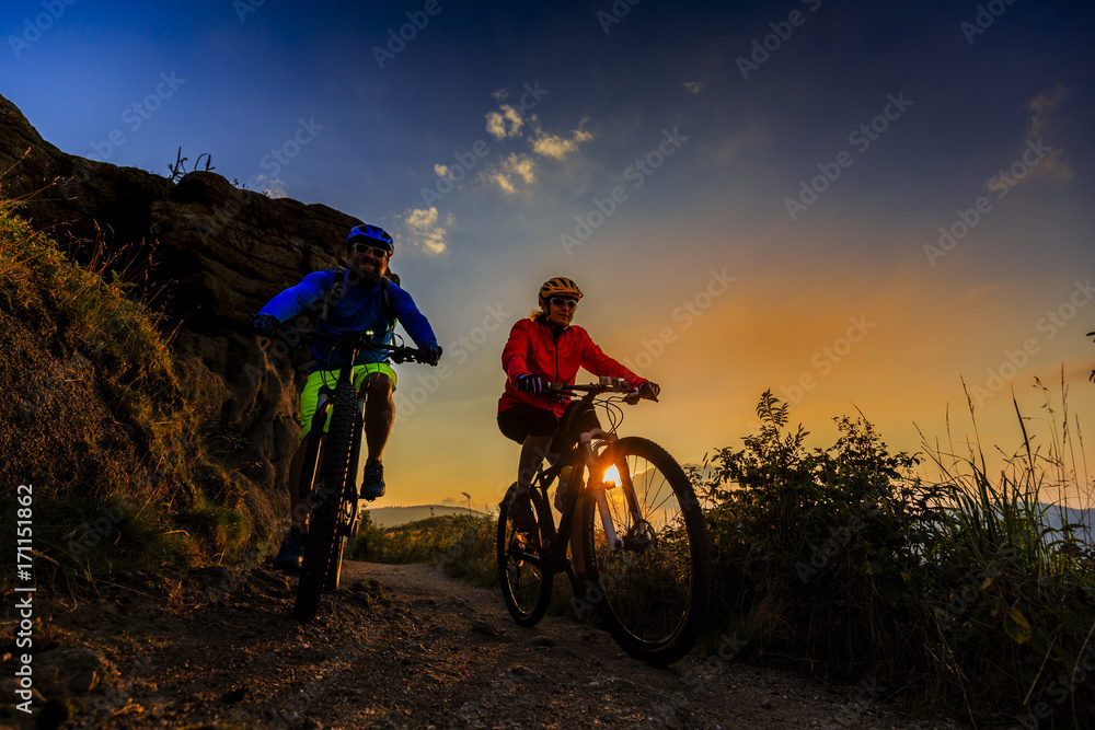 Fototapeta premium Mountain biking women and man riding on bikes at sunset mountains forest landscape. Couple cycling MTB enduro flow trail track. Outdoor sport activity.