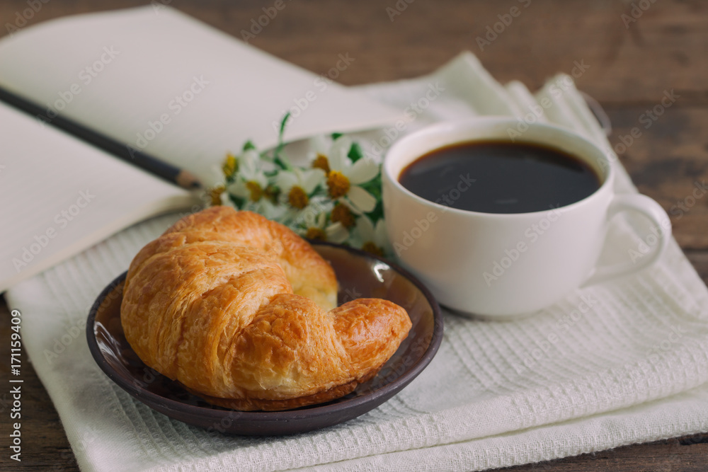 Homemade croissant served with black coffee or americano. Delicious breakfast with fresh croissant and coffee. Croissant and black coffee on wood table with open notebook and pencil with copy space.