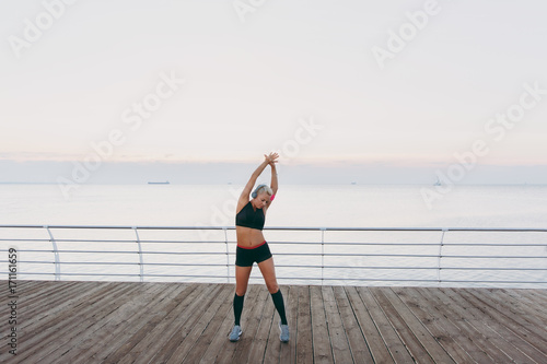 Young beautiful athletic girl with long blond hair in headphones, listening to music and making slopes at sunrise over the sea