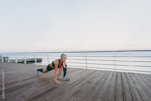 Young beautiful athletic girl with long blond hair in headphones listening to music and doing stretching at sunrise over the sea