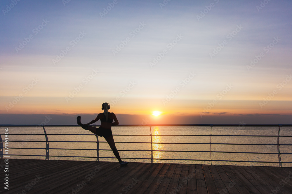 Silhouette of young beautiful athletic girl with long blond hair in headphones listening to music and doing stretching at sunrise over the sea, back view