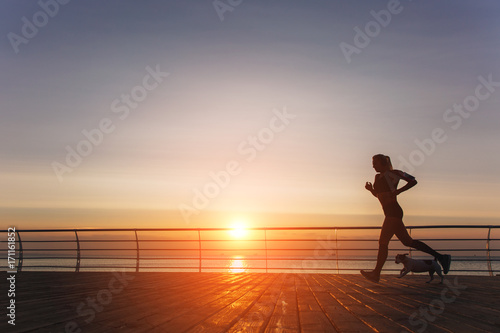 Silhouette of a young beautiful athletic girl with long blond hair in headphones that runs at dawn over the sea with her dog