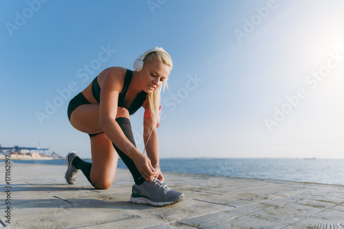 Young beautiful athletic girl with long blond hair in headphones, listening to music and lacing sneakers at dawn over the sea