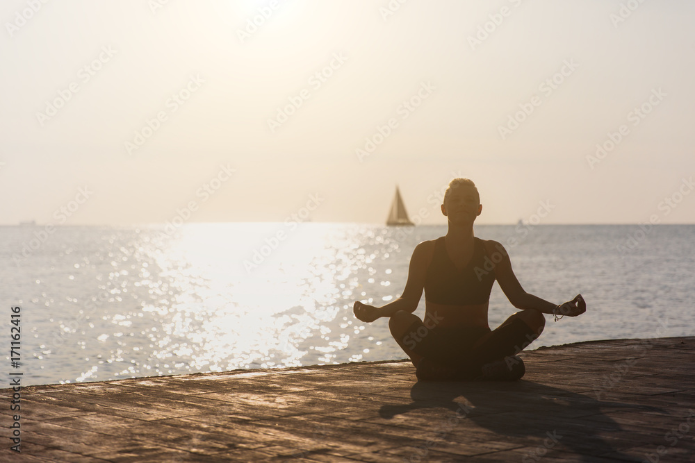 Silhouette of a young athletic girl who is engaged in yoga at sunrise on the beach with a sailboat