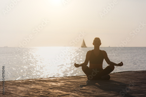 Silhouette of a young athletic girl who is engaged in yoga at sunrise on the beach with a sailboat © ViDi Studio