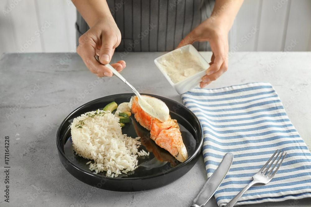 Woman pouring sauce onto plate with delicious salmon and rice in kitchen