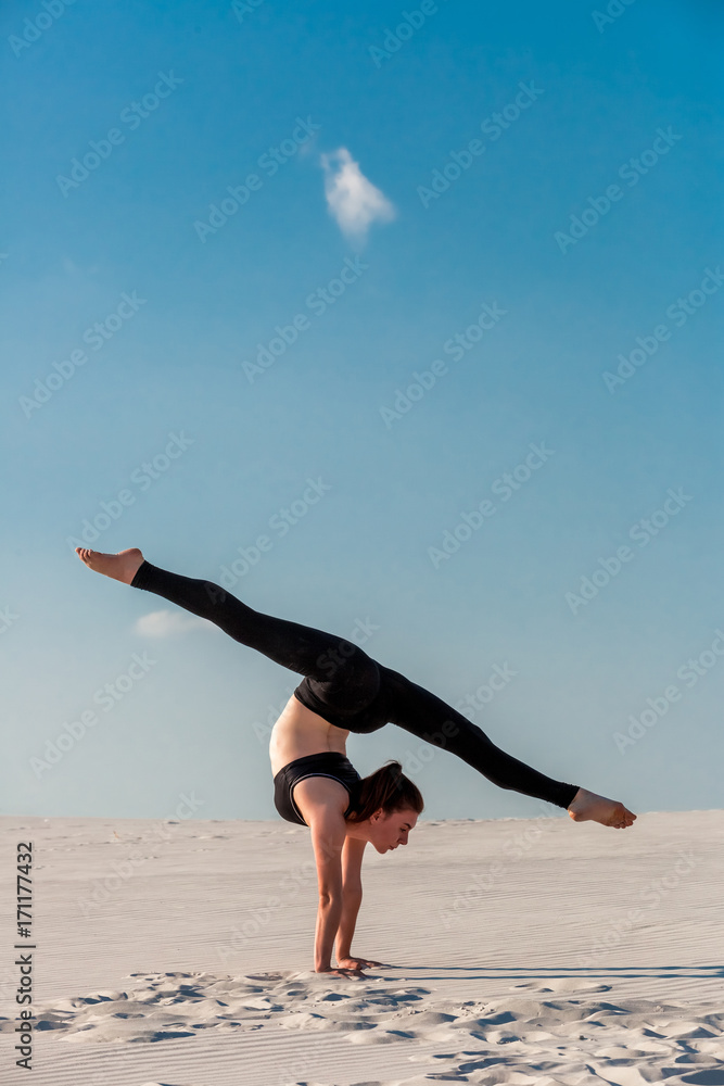 Fototapeta premium Young woman practicing handstand on beach with white sand and bright blue sky
