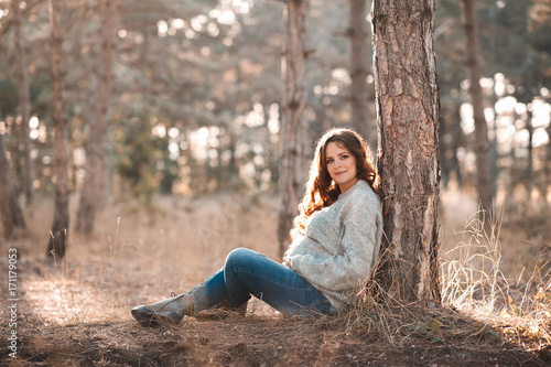 Stylish pregnant 30-35 year old wearing knitted sweater and denim pants sitting leaning on tree in woods. Looking at camera. Autumn season.