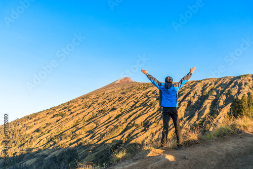 Young man hiker wear blue jacket, spreading hand, enjoy and happy with mountain top view after finished climbing at Rinjani mountain, Lombok, Indonesia.