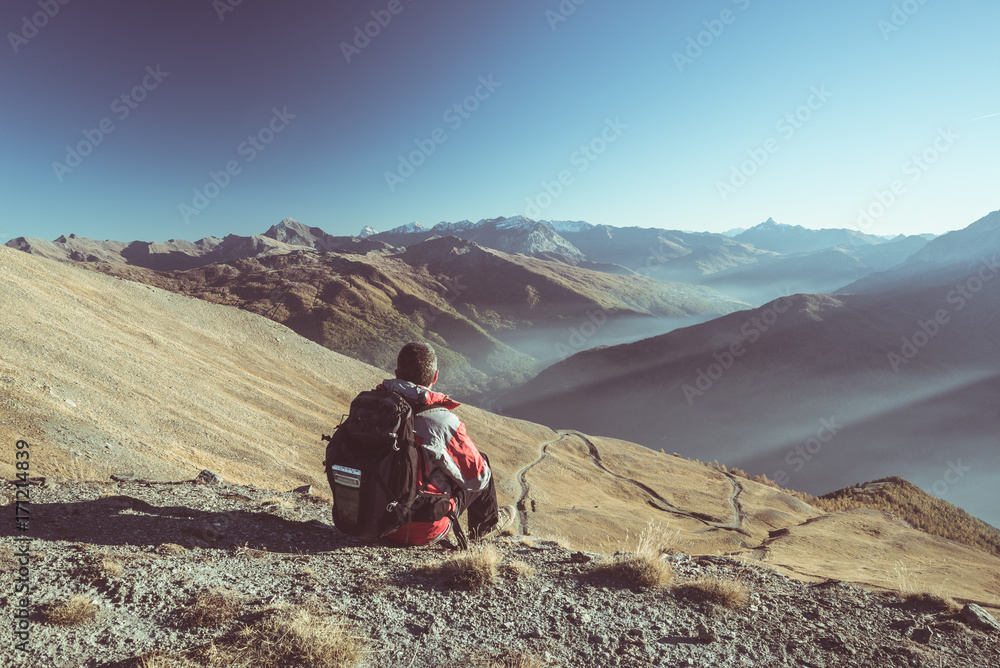 Male hiker relaxing at sunset at the mountain summit and looking at majestic panorama of the italian Alps with misty valley below. Toned image, vintage filter, split toning.