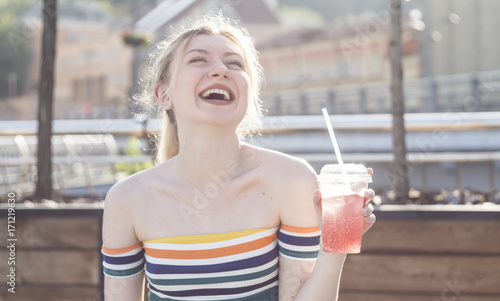 beautiful young smile blonde girl on a city street on a sunny day drinks a refreshing fruit cocktail with ice