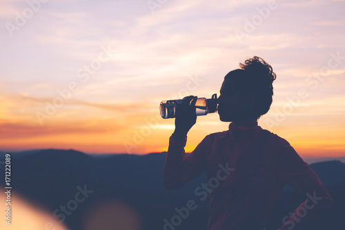 Silhouette of athletic girl drinks water after workout in the mountains at sunset. Sport tight clothes.