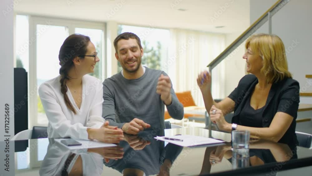 Sitting at the Table in the Living Beautiful Young Couple Signs Contract and Buys House, Real Estate Agent Gives Them House Keys. Home is Sunny and Bright. Couple is Happy, Everybody Smiles. 