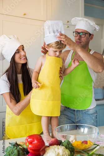 The young happy parents standing in the kitchen and dressing a little son in cook form