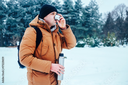 a young man in a red jacket with a backpack standing on the snow and holding a thermos and a mug of tea. Winter forest.