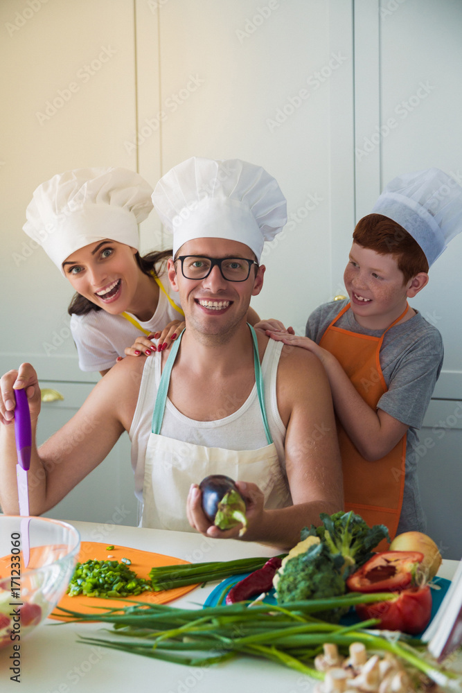 The young family in cook form in the kitchen, smiling father keeping eggplant in hand The young family in cook form in the kitchen, smiling father keeping eggplant in hand