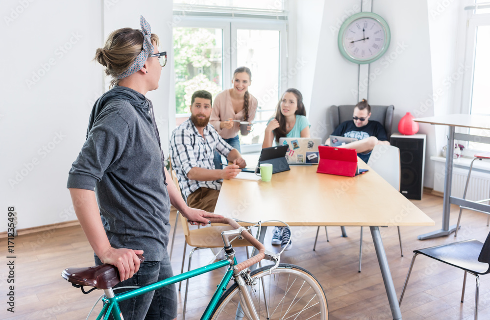 Active young woman holding a commuter bike while talking with her colleagues in the modern office of a shared co-working space for independent workers Active young woman holding a commuter bike while talking with her colleagues in the modern office of a shared co-working space for independent workers
