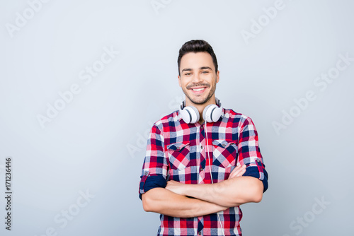 Portrait of cheerful young brunet man in casual outfit, with big white modern earphones, standing with crossed arms on the pure background