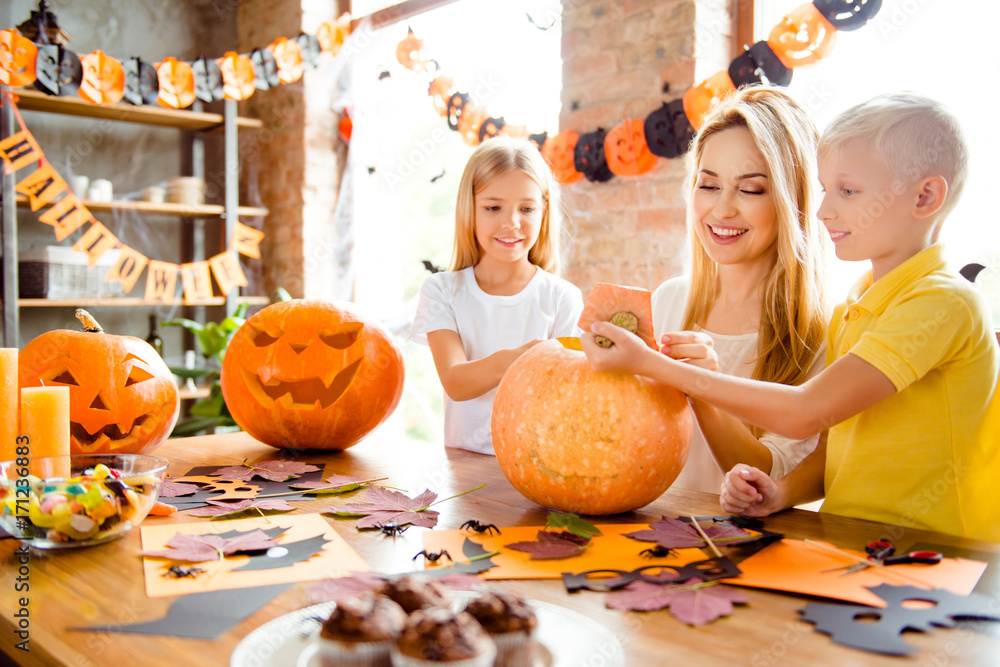 Fototapeta premium Blonde cute family of mother and two siblings making a jackolantern, boy is taking off the tail of pumpkin, yellow candles, treats, fall leaves and spiders on the table, garlands on windows