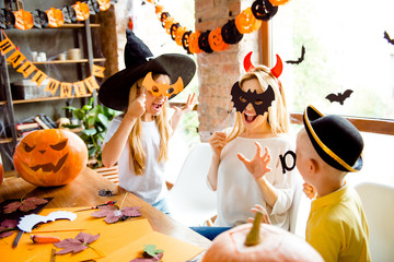 Boo! Close up shot of blond mom with two little dressed sister and brother, creepy costumes, eyewear and head wear, gesturing boo, in decorated nice loft light room indoors at home near the windows