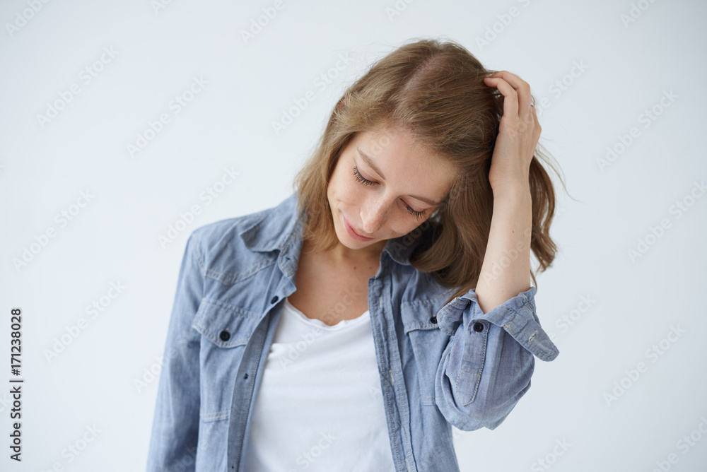 Headshot of tender and charming teenage girl with freckles looking down with shy cute smile, touching her fair hair gently, feeling confused. Genuine human emotions, feelings, reaction and attitude Headshot of tender and charming teenage girl with freckles looking down with shy cute smile, touching her fair hair gently, feeling confused. Genuine human emotions, feelings, reaction and attitude