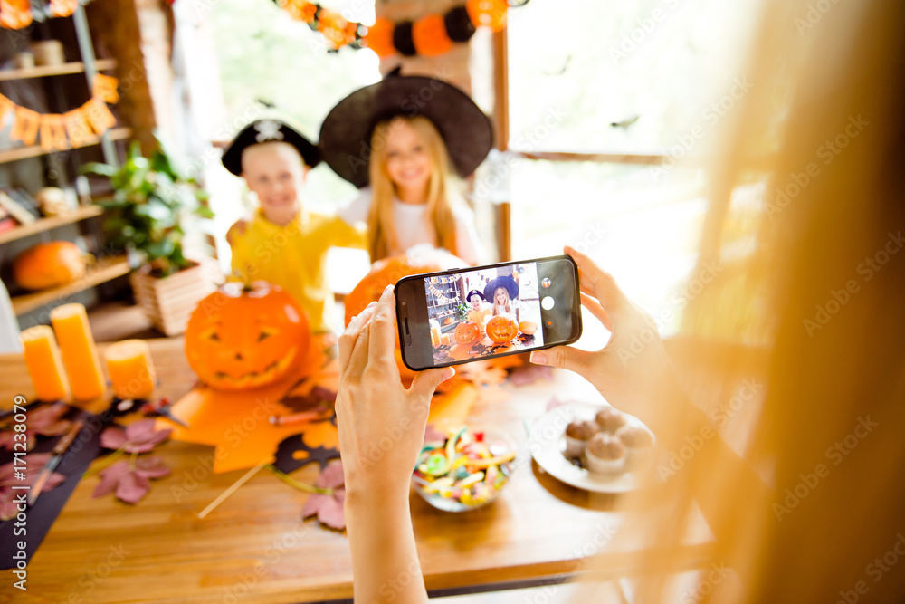 Fototapeta premium First person view, blond mommy is photographing her kids. Not clear shot of dressed in halloween headwear kids, bonding, mum is taking photo on her telephone, halloween decorations, focus of cell