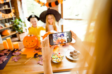 First person view, blond mommy is photographing her kids. Not clear shot of dressed in halloween headwear kids, bonding, mum is taking photo on her telephone, halloween decorations, focus of cell