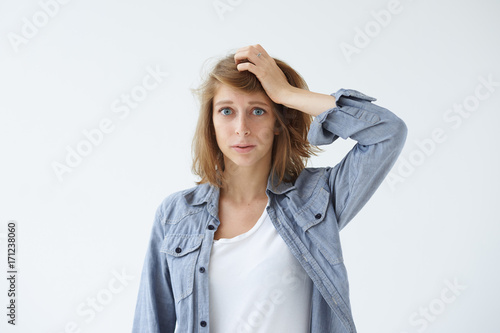 Picture of frustrated forgetful young woman looking at camera with worried puzzled expression on her face, holding hand on head as she realizes that she forgot about doing something important
