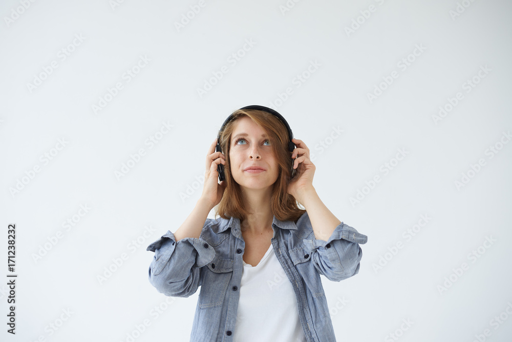 Lifestyle studio portrait of charming young female dressed in casual clothing standing at blank wall with headphones, lsitening to music tracks or audiobook, searching for inspiration, looking up Lifestyle studio portrait of charming young female dressed in casual clothing standing at blank wall with headphones, lsitening to music tracks or audiobook, searching for inspiration, looking up