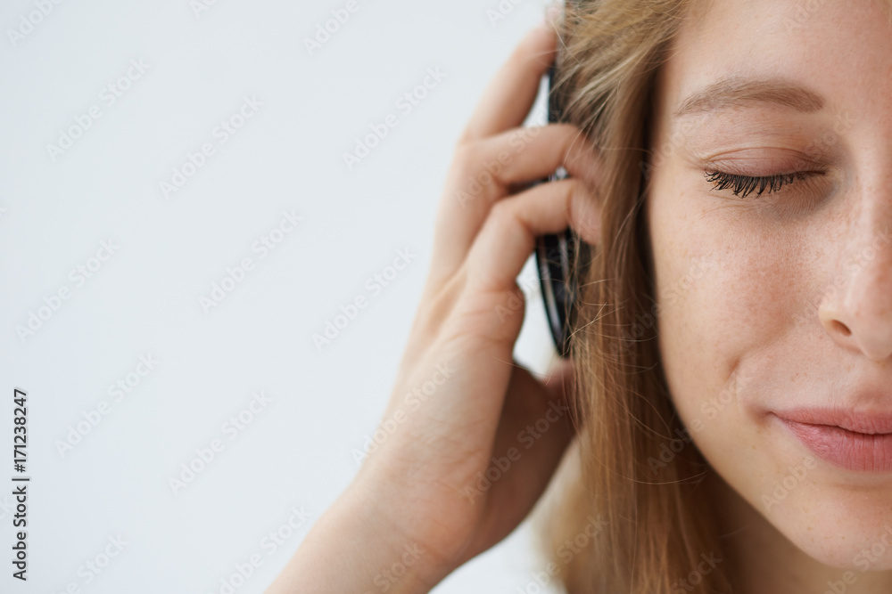Cropped shot of half face of positive joyful teenage girl wearing headphones, listening to relaxing music, meditating with closed eyes against white wall background with copy space for your text Cropped shot of half face of positive joyful teenage girl wearing headphones, listening to relaxing music, meditating with closed eyes against white wall background with copy space for your text