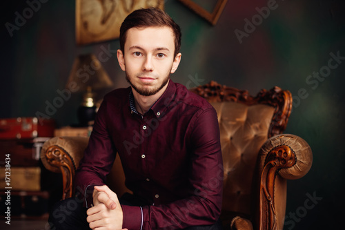 stylish young man in burgundy shirt sitting in luxury armchair a