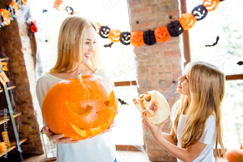 Adorable small blonde girl is showing her mom a scull decoration for a halloween party, pretty mom is with big carved pumpkin, in room with garlands, bats and spiders on the window, loft style
