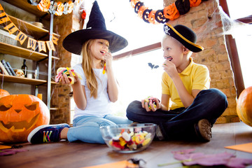Treat or trick! Low angle shot of enjoying junior blond kids in carnival outfits, caps, with colorful treats, eating them, sitting on the floor, near the window in halloween decorated room at home