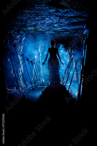Silhouette of a mysterious witch saying the spell and stretching her hands to us, with clouds of smoke around her, and dungeon bridge on the background