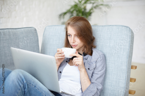 Casually dressed female college student enjoying coffee or tea while watching film online on laptop computer at weekend. Pretty girl watching video blog using wifi on notebook pc, sitting on sofa