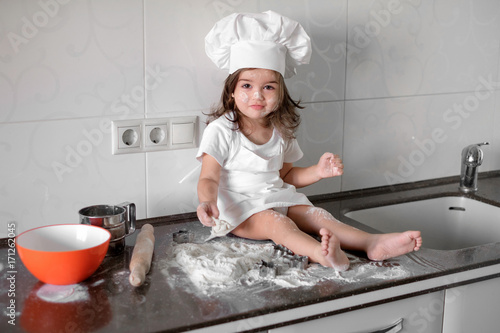Cute little girl in apron and chef hat is kneading the dough and smiling while baking photo