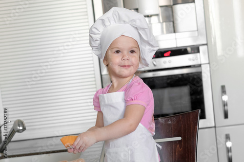 Cute little girl in apron and chef hat is kneading the dough and smiling while baking photo