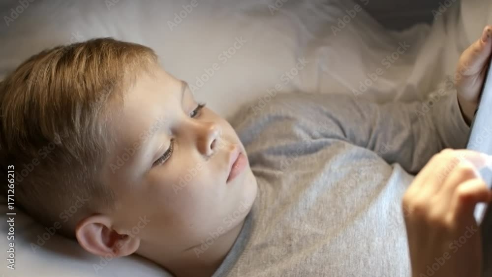 PAN with top view of little boy with blond hair lying in bed and reading digital book on tablet PAN with top view of little boy with blond hair lying in bed and reading digital book on tablet