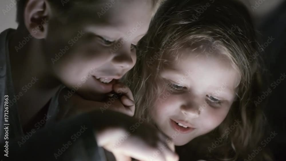 PAN with close up of laughing girl and boy playing on tablet in dark bedroom PAN with close up of laughing girl and boy playing on tablet in dark bedroom
